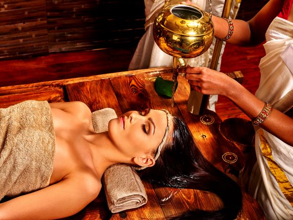 krásná žena ležící na dřevěném stole realaxuje a na hlavu jí teče teplý bylinkový olej z měděné nádoby. To je součást panchakarmi.